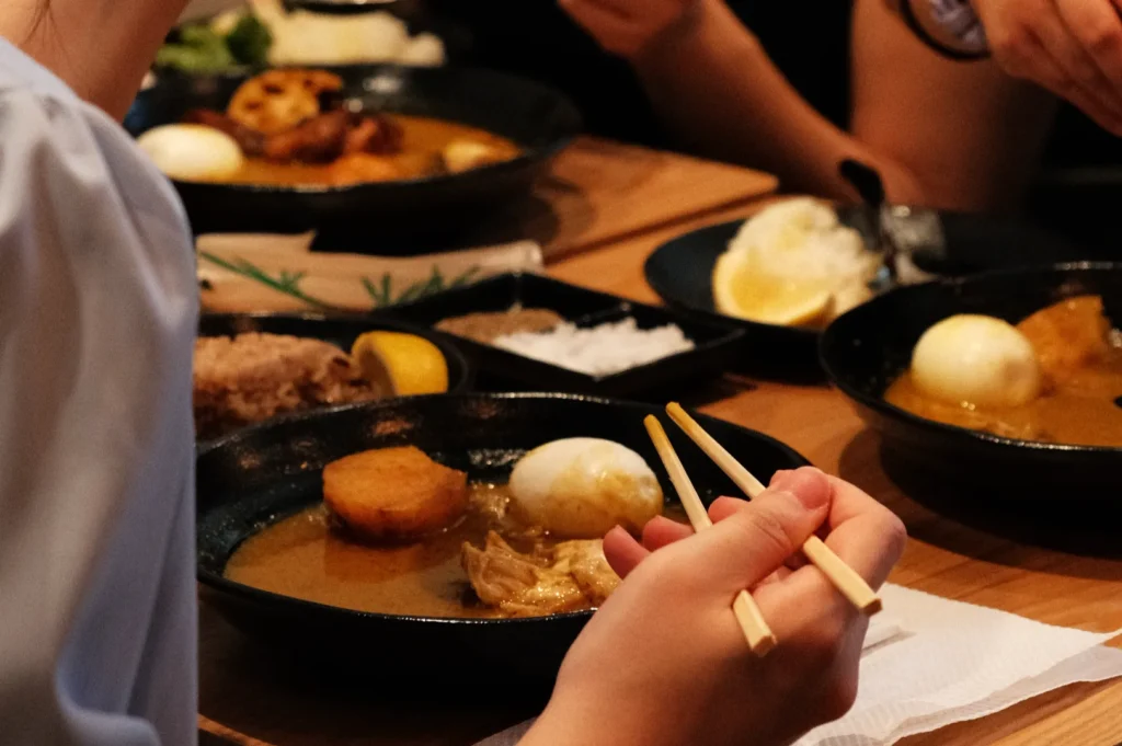 A close-up of hands using chopsticks to eat a dish, with a bowl of food including a soft-boiled egg, vegetables, and broth on a wooden table, surrounded by other bowls and dishes.