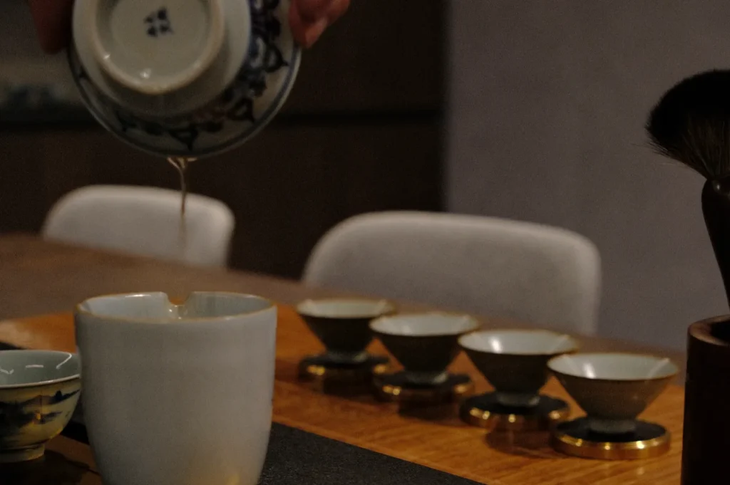 A hand pours liquid from a decorative cup into a plain mug, with four small teacups in the background on a wooden table. The scene feels calm and intimate.