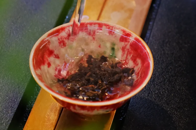 A red ceramic bowl with patterns being filled with hot water, infusing loose tea leaves. Steam rises, creating a warm, soothing atmosphere.