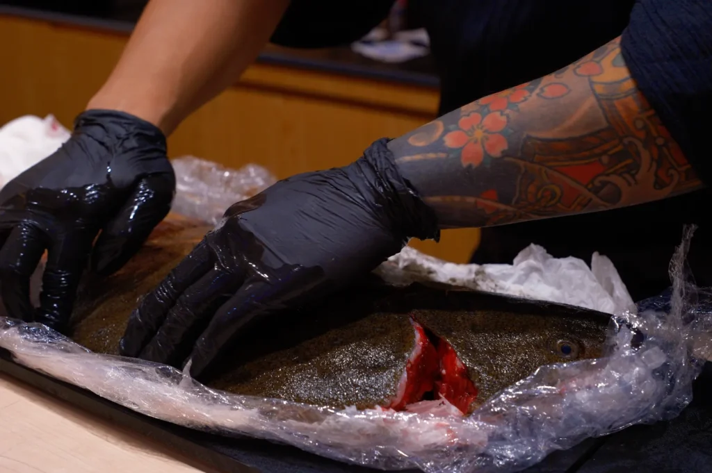 Tattooed arms with black gloves preparing a large fish on a table. The fish is wrapped in plastic, with focus on the detailed tattoos and careful handling.