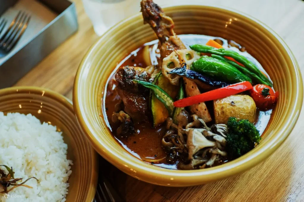 Bowl of vibrant curry with chicken drumstick, mixed vegetables, and mushrooms. Served with a side of white rice on a wooden table, creating a cozy dining atmosphere.