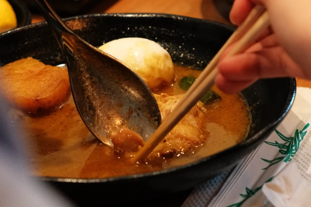 A close-up of a bowl of ramen with a person using chopsticks to pick up a piece of meat from the broth, which also contains a boiled egg and other ingredients.