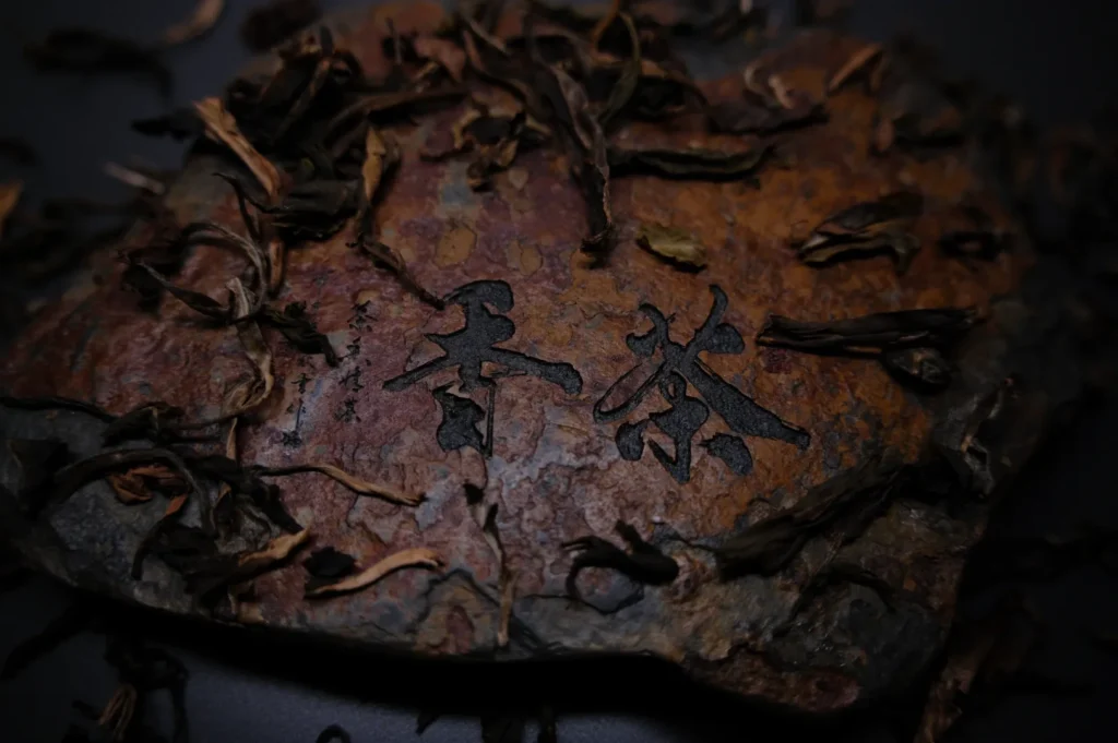 A dark, rustic piece of tea brick with Chinese characters etched on its surface, surrounded by scattered, dried tea leaves on a black background.