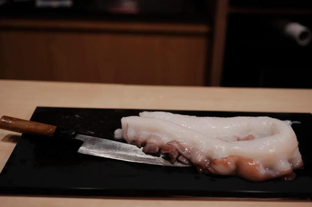 Fresh octopus tentacle on a black tray with a wooden-handled knife, placed on a light wooden counter. The scene is calm and culinary-focused.