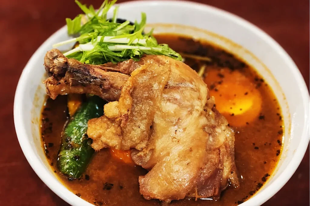 A bowl of rich, dark curry soup topped with a crispy fried chicken leg, vibrant green herbs, and a whole green chili. The setting is warm and appetizing.