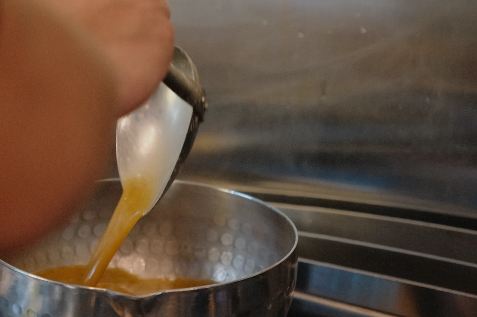 A person's hand pouring a yellowish liquid from a ladle into a metal bowl.