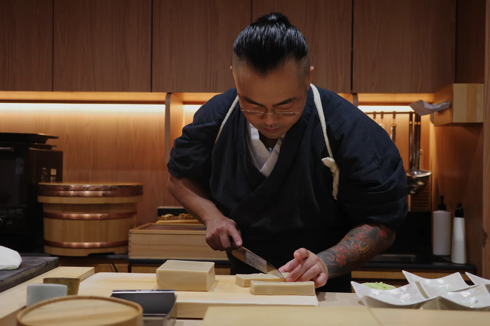 Chef Masa in a dark outfit is skillfully preparing food, using a knife to cut rectangular blocks of an ingredient on a wooden countertop.