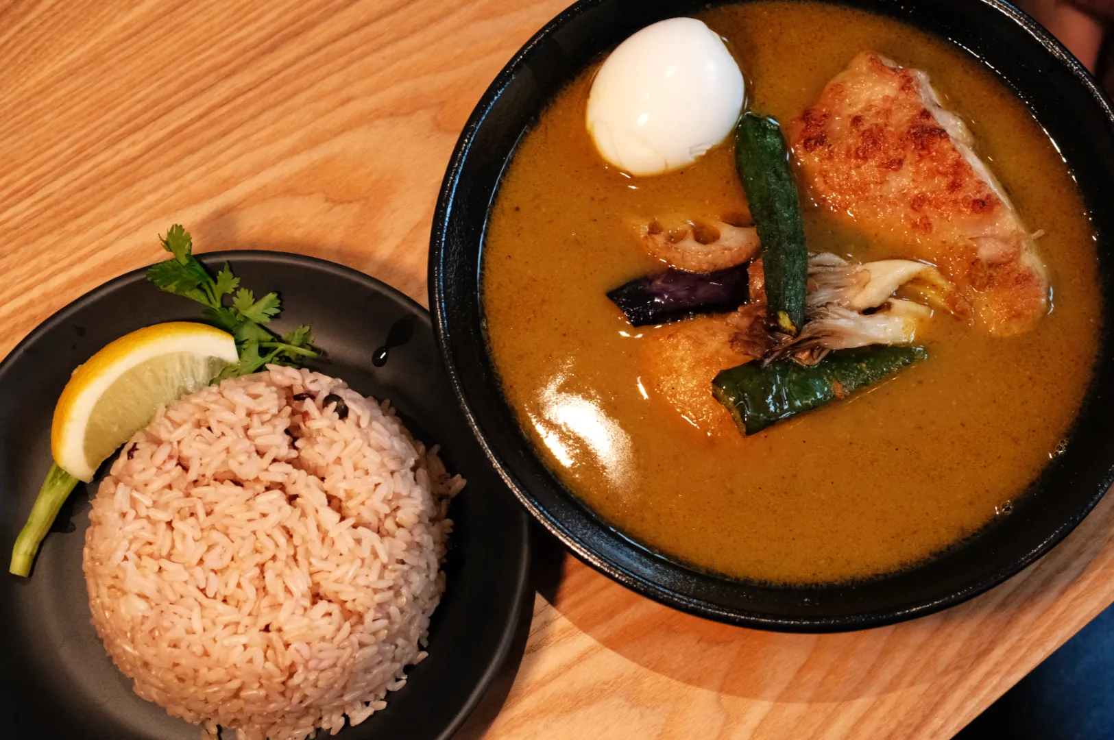 A bowl of soup curry with vegetables and chicken, accompanied by a boiled egg and a side of brown rice with a lemon wedge and a sprig of cilantro.