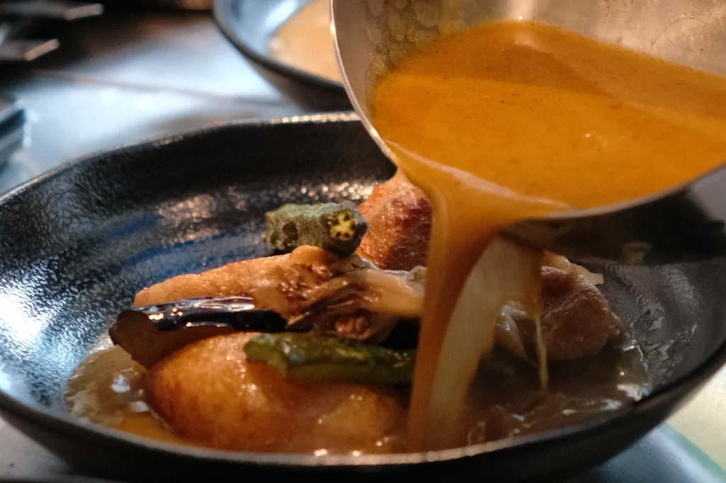 A chef pouring sauce over a dish featuring vegetables and dumplings in a dark bowl.