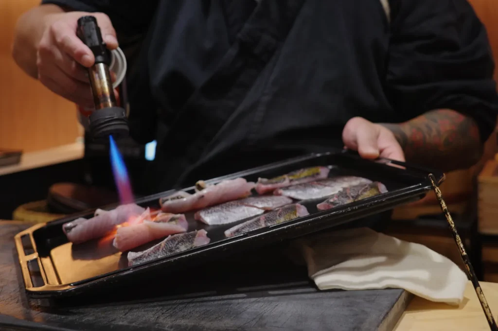 Chef Masa in a black traditional uniform using a kitchen torch to sear fish fillets on a tray.