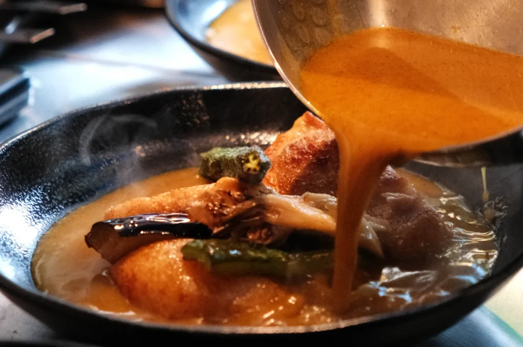 A close-up of a bowl containing a hot, steaming soup curry with vegetables, while broth is being poured over it.