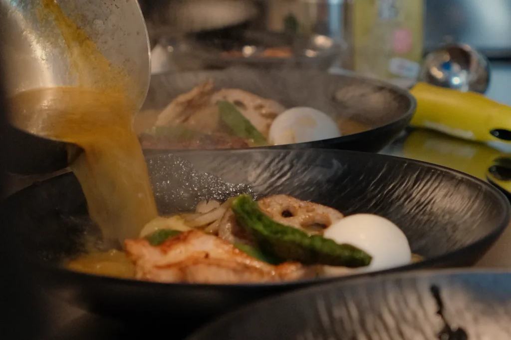 A steaming bowl of ramen being served, with noodles, meat, vegetables, and an egg, in a black bowl.
