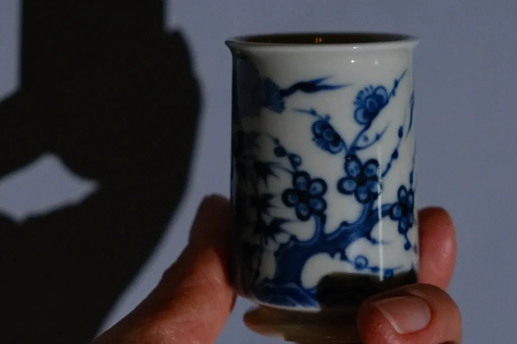A hand holding a small ceramic cup with blue floral designs, casting a shadow on a surface.