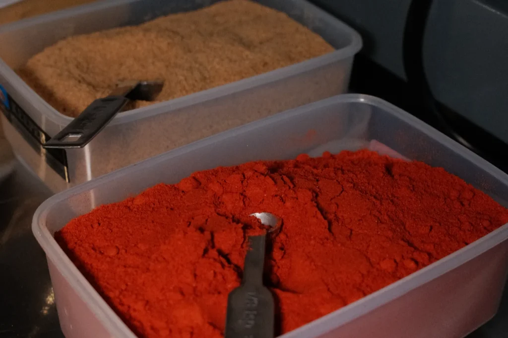 Two containers of spices, one filled with red powder and the other with brown granules, each with a measuring spoon.