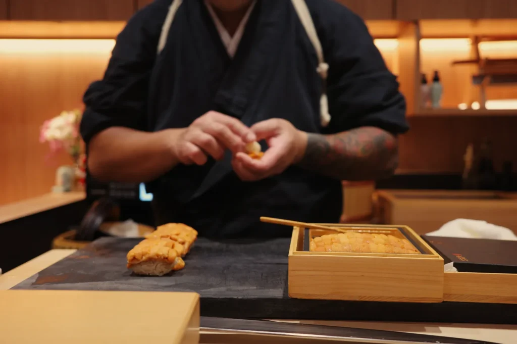 Chef Masa preparing sushi in a wooden setting, with sushi pieces and a bamboo box on the counter.