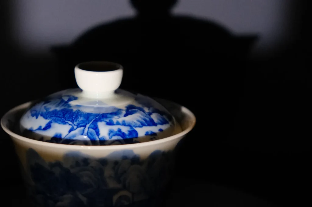 A decorative bowl with a lid, featuring blue and white floral patterns, is set against a dark background, casting a shadow.