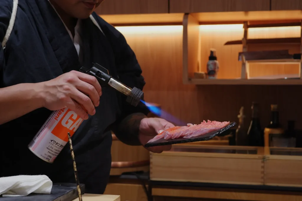 Chef Masa wearing a black apron is using a kitchen torch to sear slices of sushi on a black plate in a well-lit kitchen.