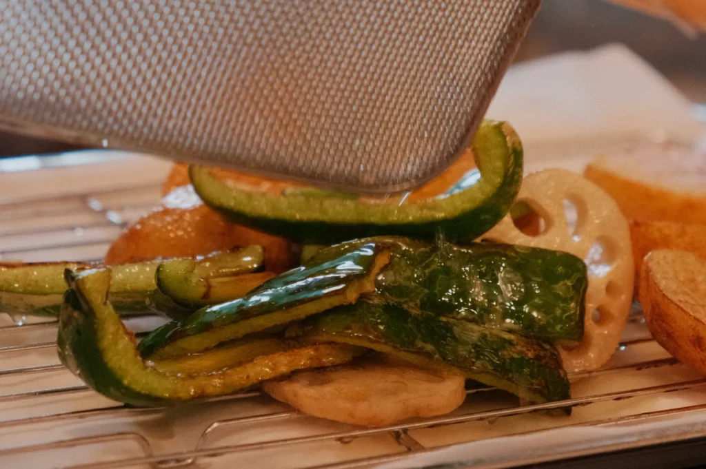 A metal mesh spatula hovering over a plate of flash fried vegetables, including zucchini, peppers, and other assorted items, sitting on a wire rack.