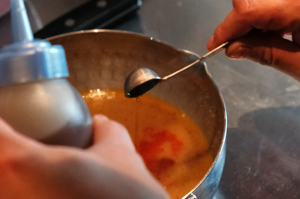 A person is holding a small measuring spoon above a mixing bowl while pouring from a squeeze bottle.