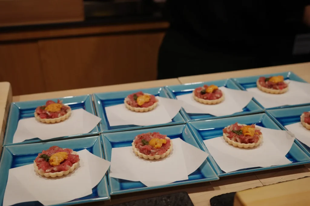 A display of small, circular tartlets topped with a mixture of diced raw fish and garnishes, presented on blue plates.