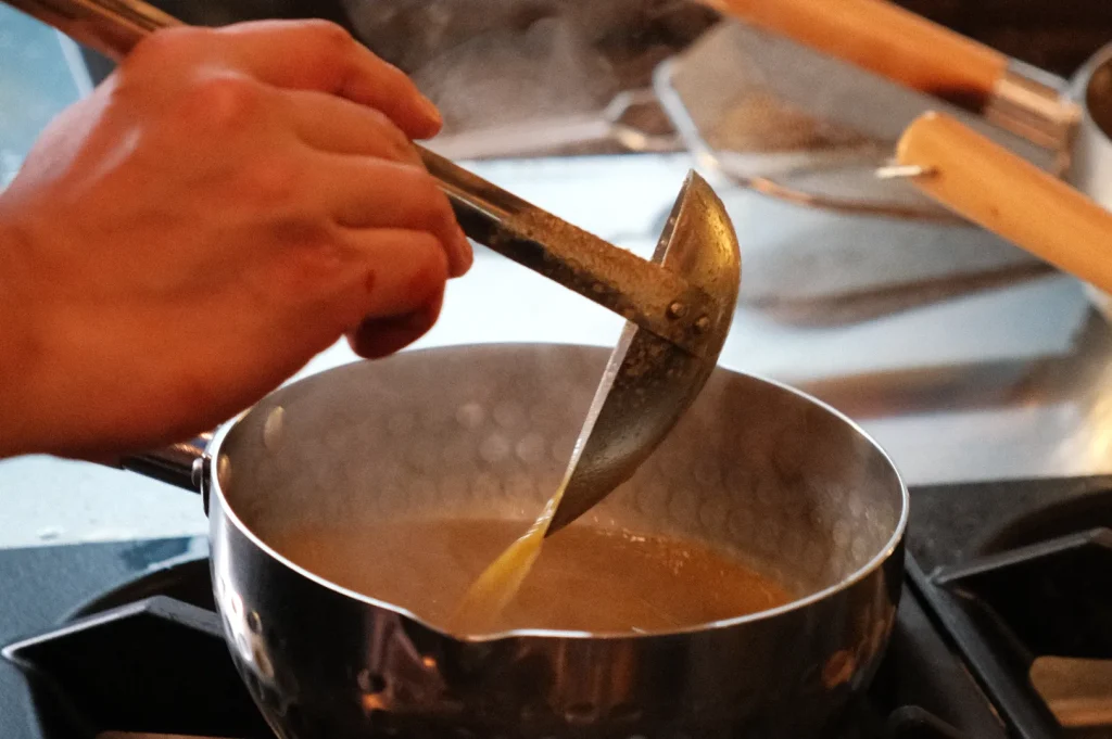 A person's hand using a tool to stir a pot on a stove. The pot contains a simmering liquid.