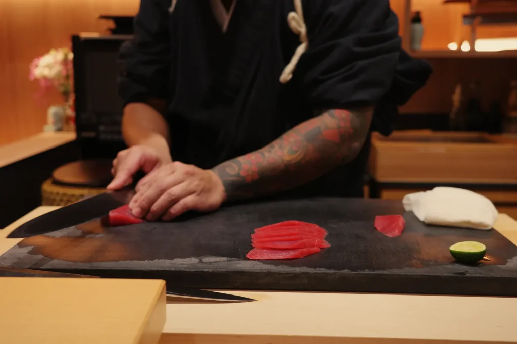 Chef Masa in a traditional setting is preparing sushi, using a sharp knife to slice fresh fish on a dark stone surface, with a small lime and rice nearby.