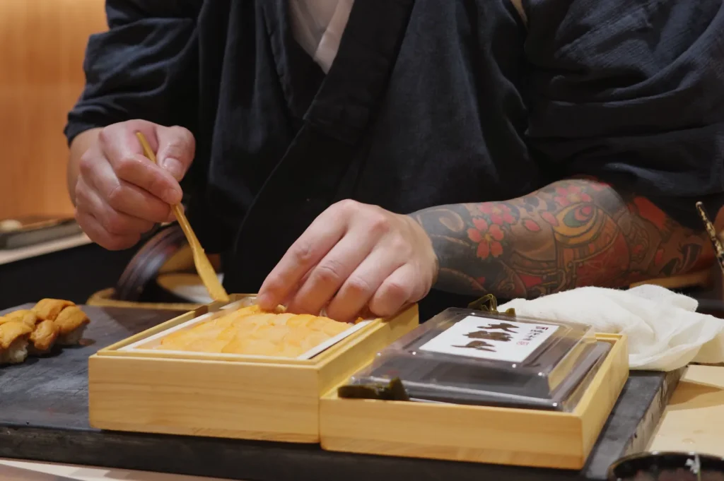 A person with tattooed arms assembling food in a wooden box, using a small utensil, in a culinary setting.