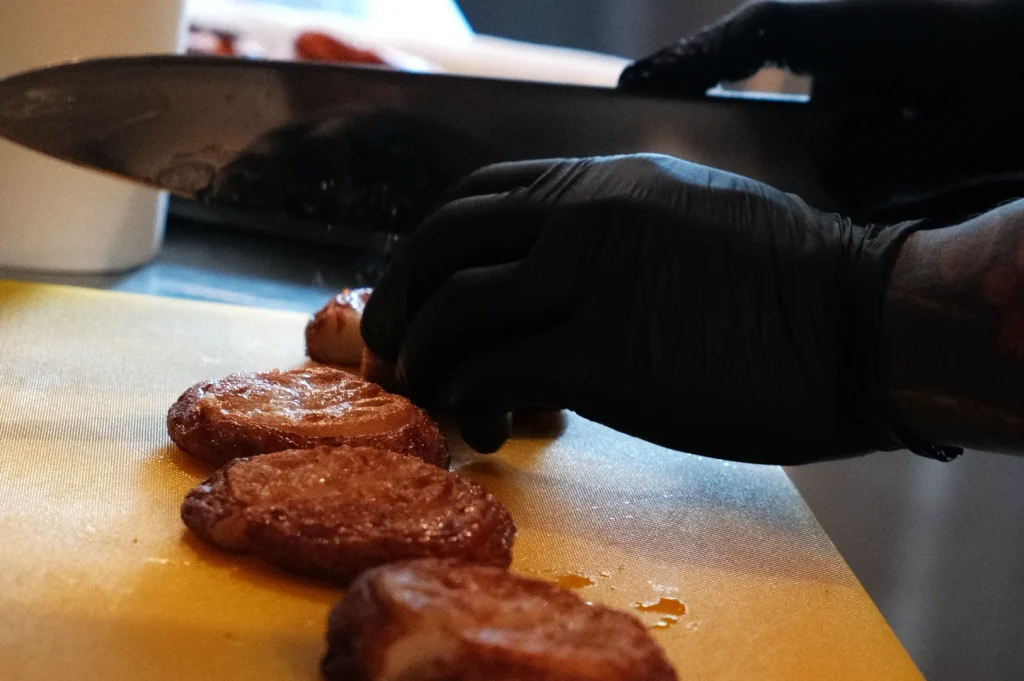 A person wearing black gloves is slicing cooked food items on a yellow cutting board.