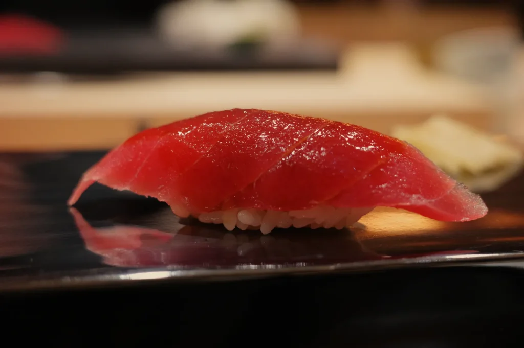 A close-up of a piece of sushi, specifically a slice of tuna sashimi, presented on a black plate.