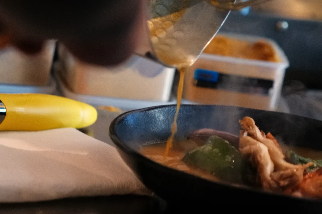 A close-up of a person pouring a yellow liquid into a black bowl containing food with visible green bell peppers and chicken.