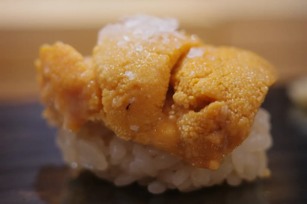 A close-up of sushi topped with a bright orange piece of sea urchin (uni) on a bed of rice, sprinkled with a pinch of salt.