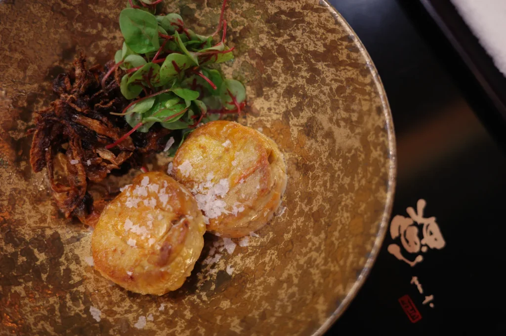 A decorative plate featuring two golden-brown savory pancakes, topped with a sprinkle of salt and served alongside a small mound of fried shallots and fresh microgreens.