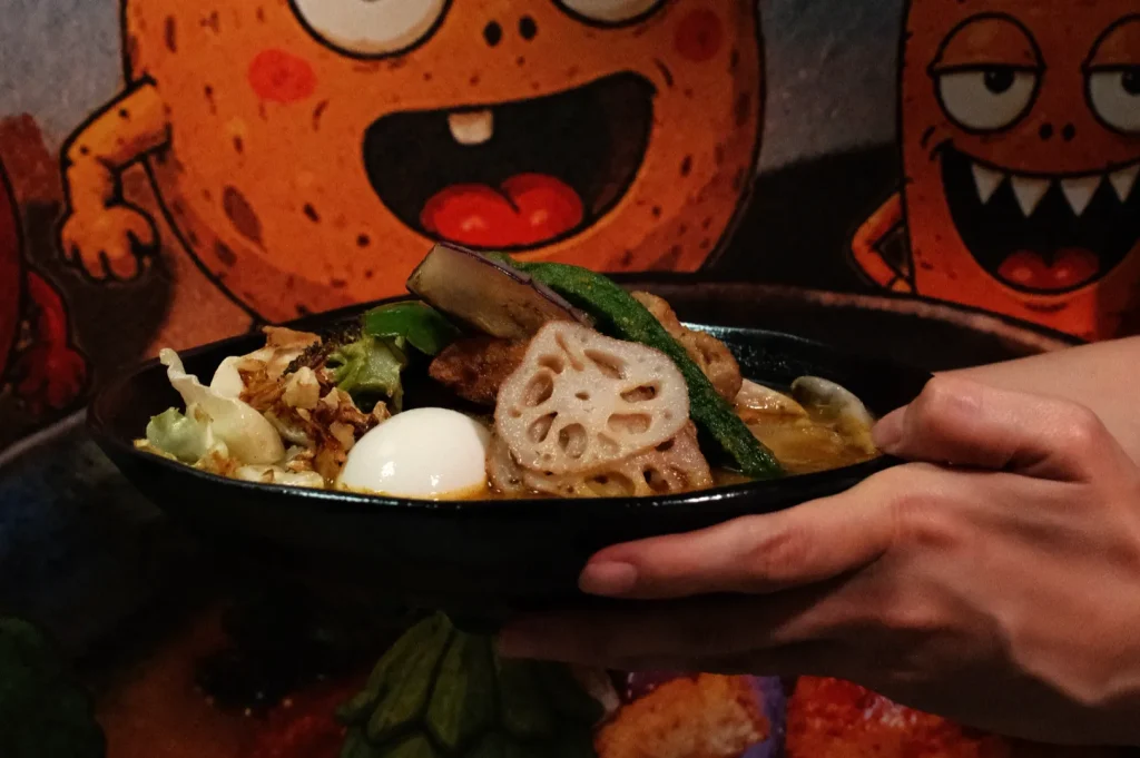 A person holding a bowl of ramen topped with various ingredients, including lotus root, vegetables, and an egg, with a colorful mural in the background.