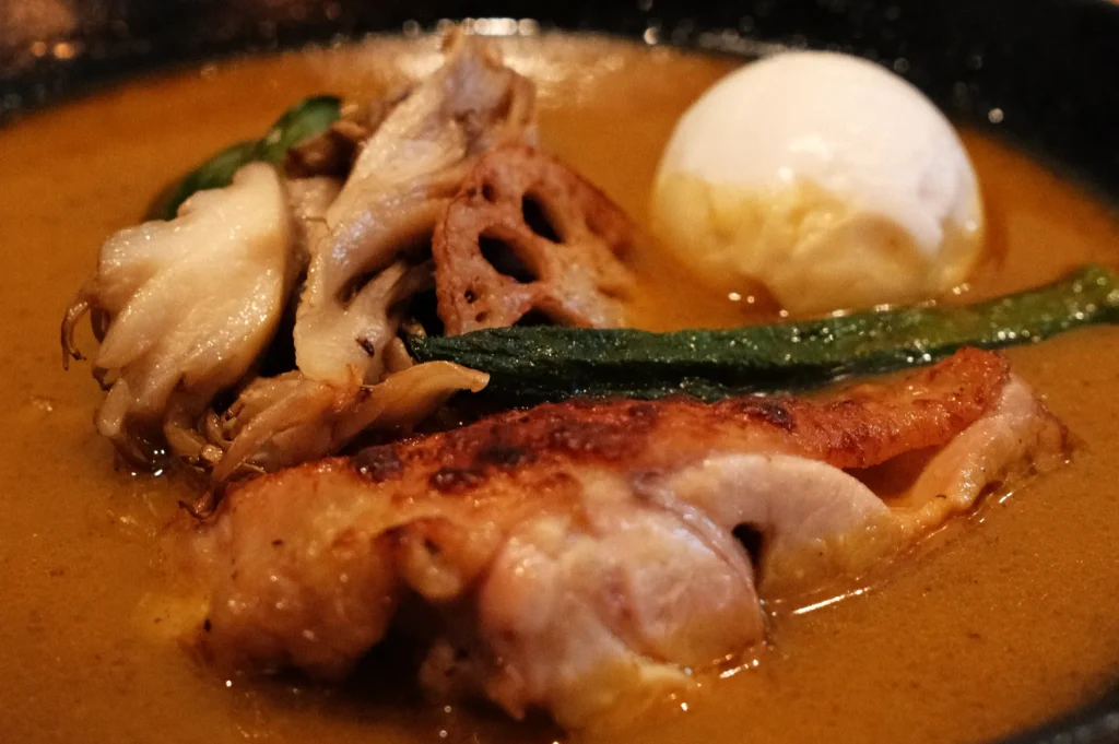 A bowl of soup curry featuring a soft-boiled egg, chicken, mushrooms, lotus root, and green vegetables in a rich broth.