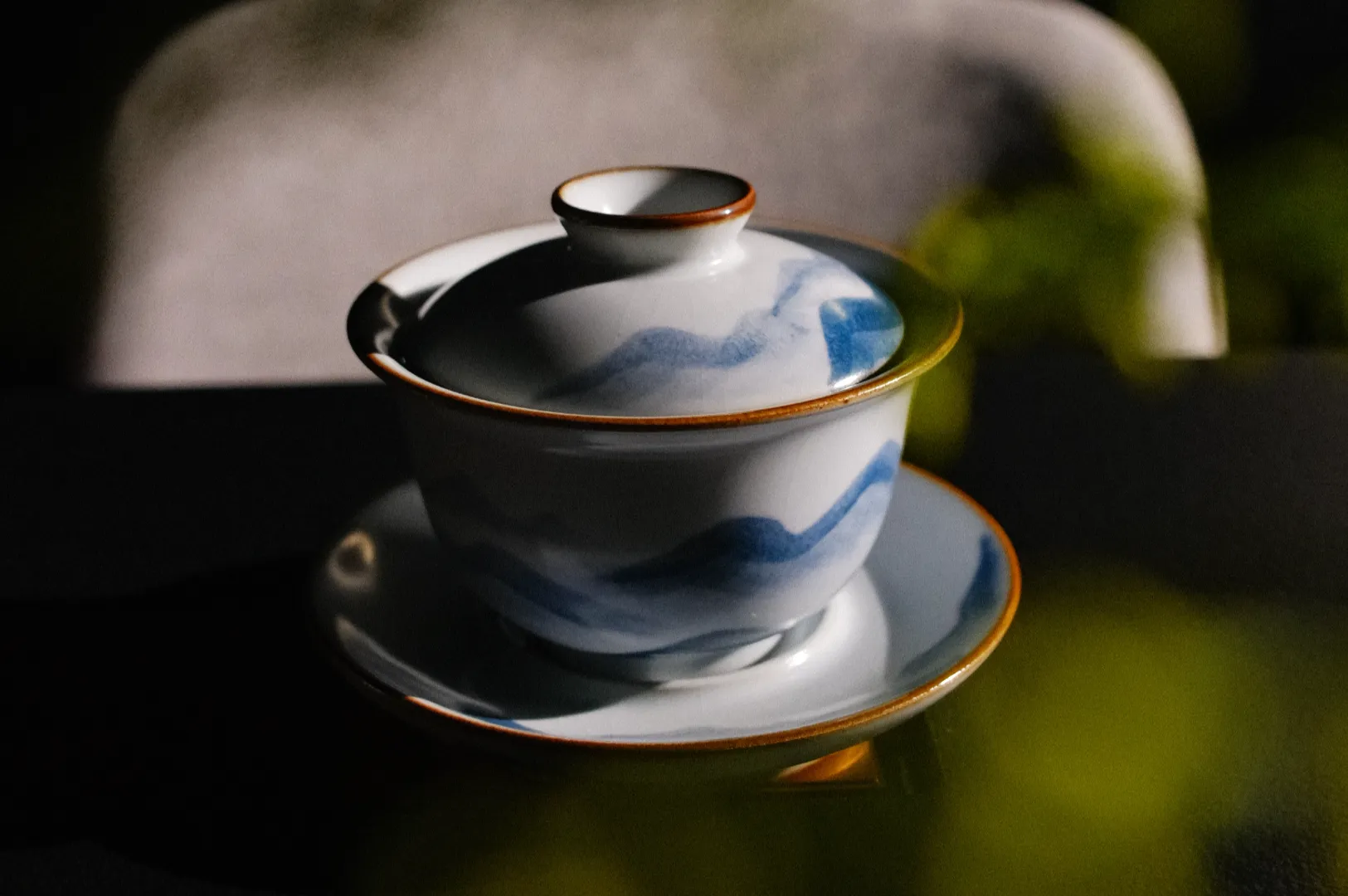 A decorative teacup and saucer with a blue wave pattern and a lid, placed on a dark surface with soft lighting and a blurred background.