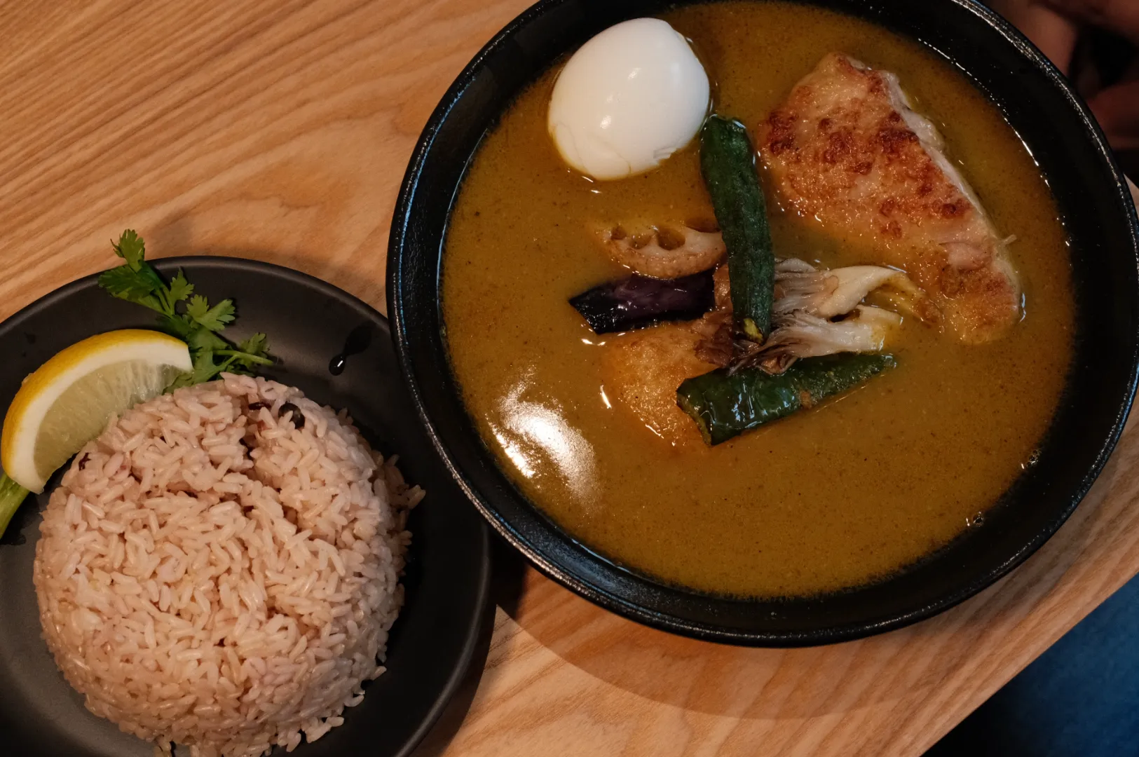 A bowl of curry soup with chicken, green vegetables, and a soft-boiled egg, served alongside a plate of brown rice and a lemon wedge.