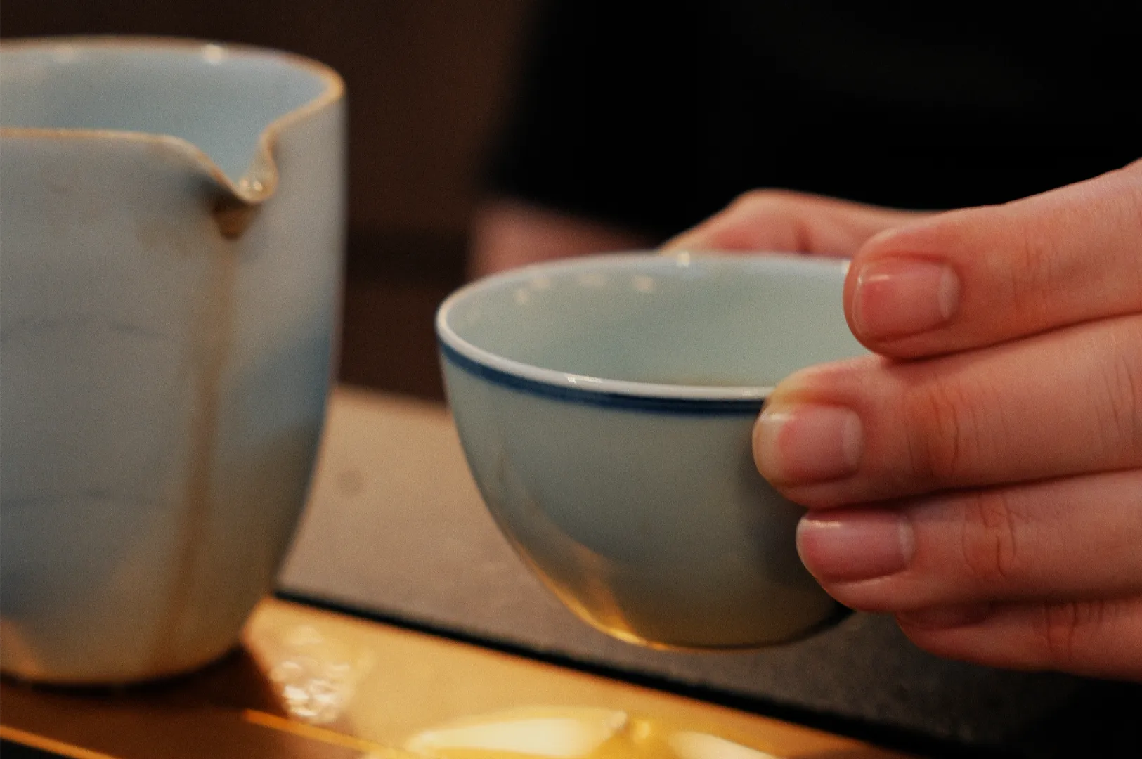 Hand holding a blue ceramic teacup beside a matching pitcher on a wooden table, creating a warm, intimate tea-serving scene.