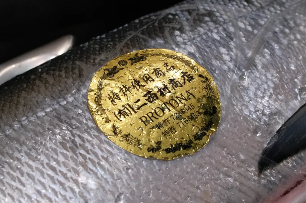 A close-up of a fish with a shiny gold label attached to its skin. The label has Chinese characters and the word "PROTONA.”