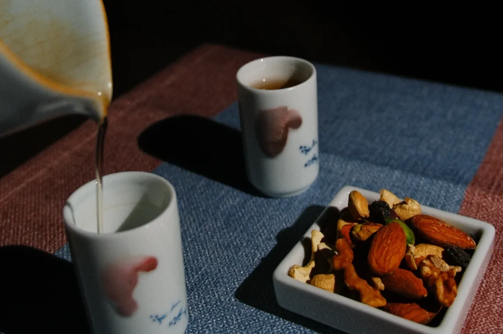 A table setting with two small cups, one being filled with a liquid, and a square bowl of mixed nuts and dried fruits.