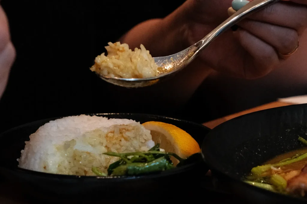 A person holding a spoonful of rice above a bowl of rice with herbs and a lemon slice.