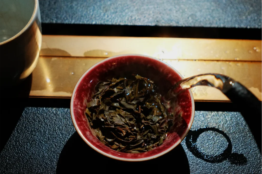 A red bowl filled with dark, wet tea leaves sits in sunlight on a black surface, beside a spoon and partial metal cup, creating a rustic mood.