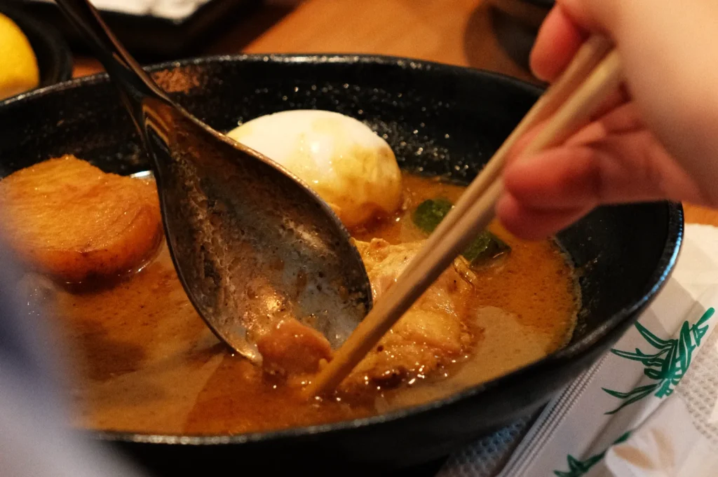 A hand holding chopsticks scooping food from a bowl of ramen, which contains slices of meat, an egg, and broth.