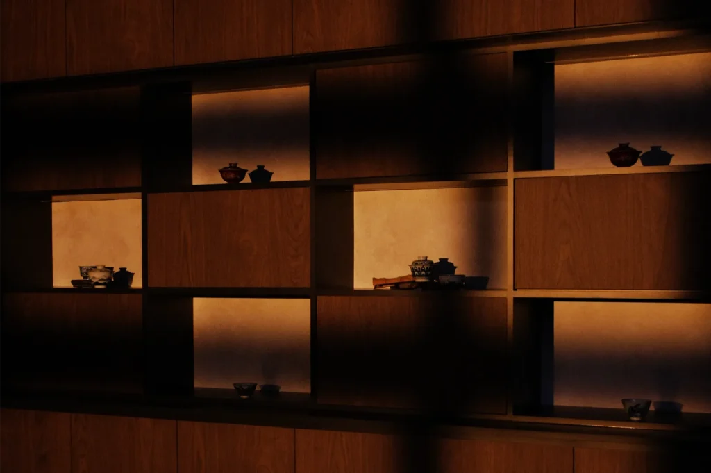 Moody, warmly lit wooden shelving displays small dark ceramic bowls and ornaments in recessed compartments, creating a quiet, minimalist atmosphere.