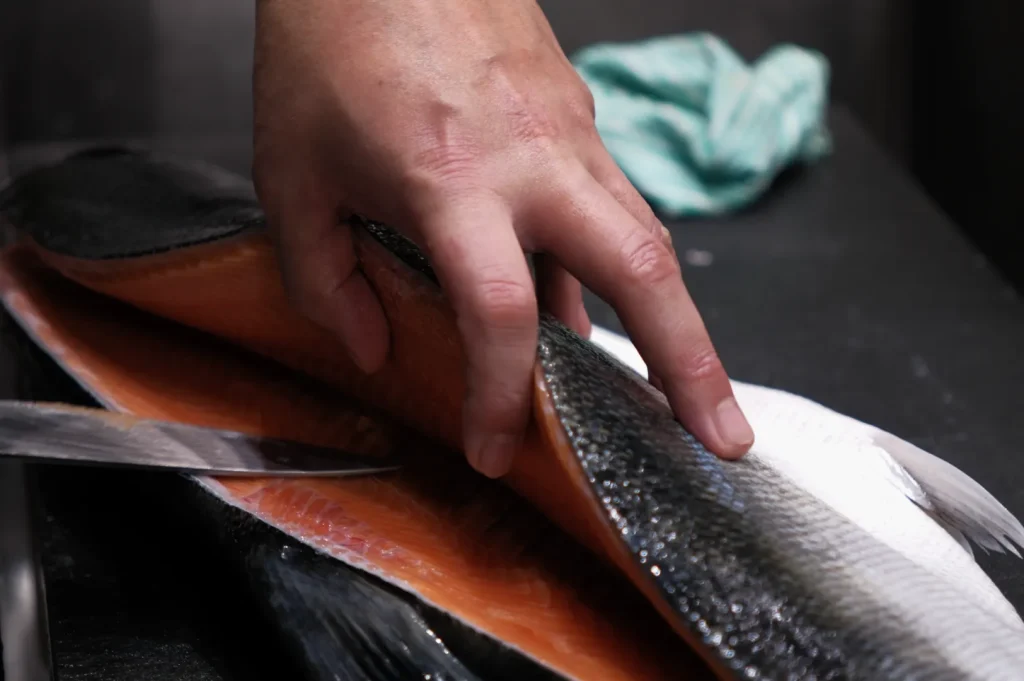A hand holding a piece of fish while a knife is positioned nearby.