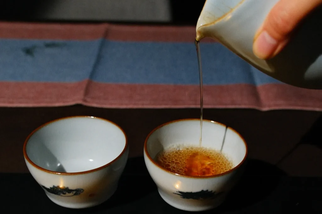 A hand is pouring tea from a teapot into two empty cups on a dark surface with a cloth in the background.