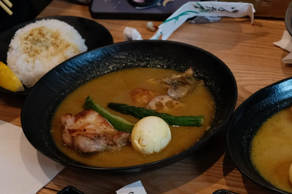 A bowl of soup with pieces of meat, half a boiled egg, and green vegetables, accompanied by a serving of rice on the side.
