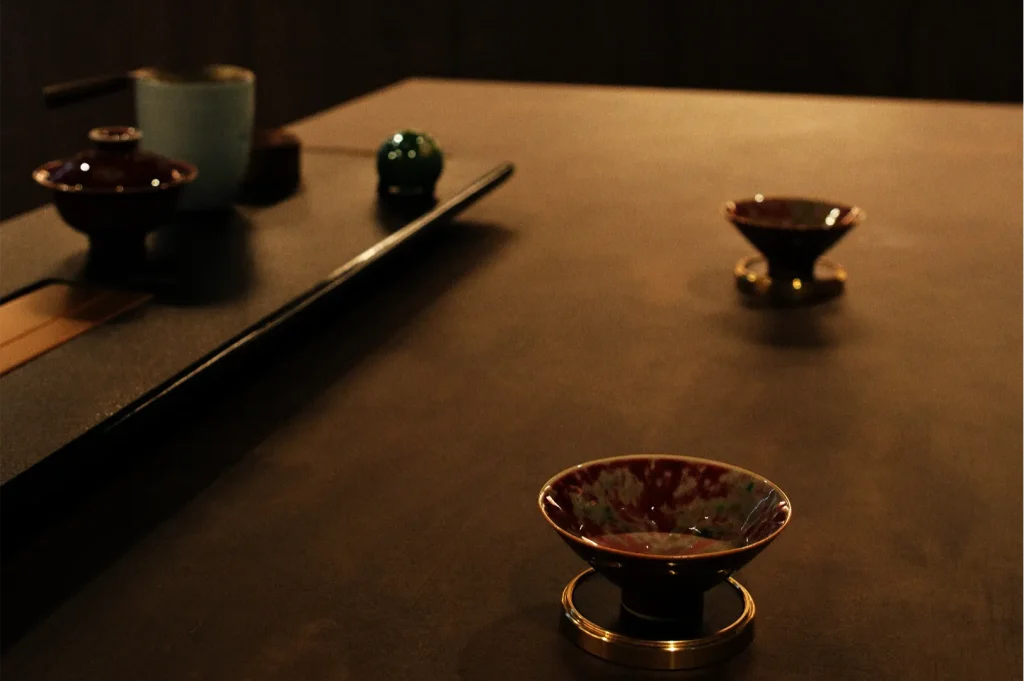 Warmly lit tabletop with Japanese tea cups, a dark serving tray, bamboo whisk, and green ornament, creating a calm, intimate mood.