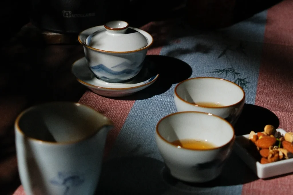 A tea setup with a teapot, multiple small tea cups, and a plate with assorted snacks, arranged on a textured cloth.
