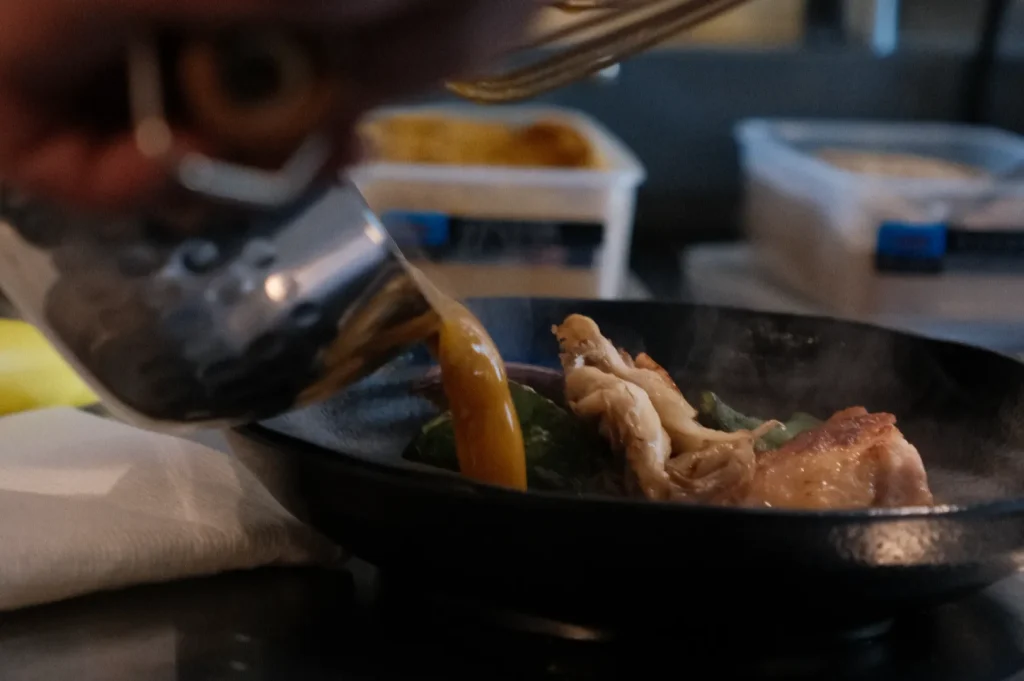 A close-up of a hand pouring sauce onto a sizzling dish in a frying pan, with vegetables and meat visible. Steam is rising from the pan.
