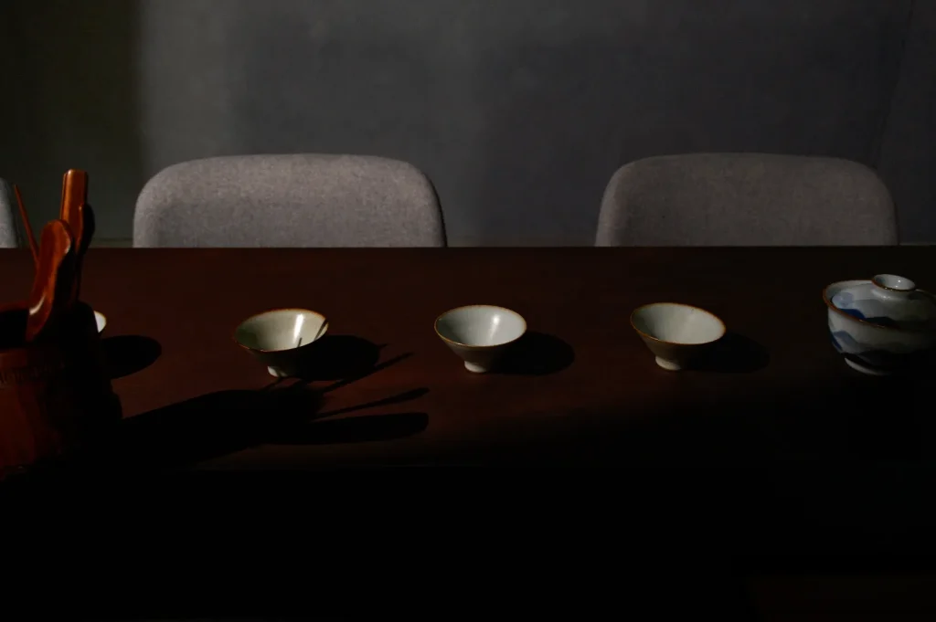 Moody still life of three small white tea cups on a dark wooden table, with a teapot at right, empty chairs behind, and deep shadows.